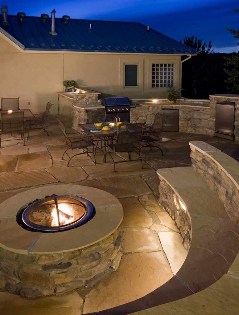 Backyard outdoor kitchen at dusk with lighting and a fire pit.