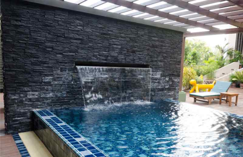 Swimming pool with stylish gray stone veneer on one wall with a fountain spilling into the pool