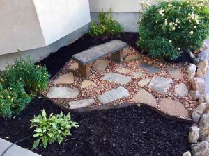 A newly landscaped flowerbed with flat rocks, orange pebbles, a rose bush, Snapdragon flowers, dark mulch, and a stone bench.