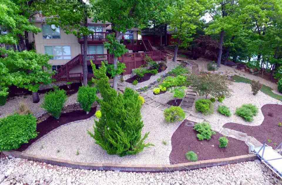 Aerial view of a low-maintenance lakeside landscape in Missouri featuring light-colored gravel ground cover, winding stone pathways, established shrubs, and mature trees around a home with a large wooden deck.